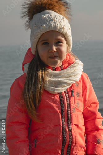 a little girl in a winter suit plays on the seashore, a girl poses on the seashore, a little girl in a hat with a bumbon plays near the sea photo