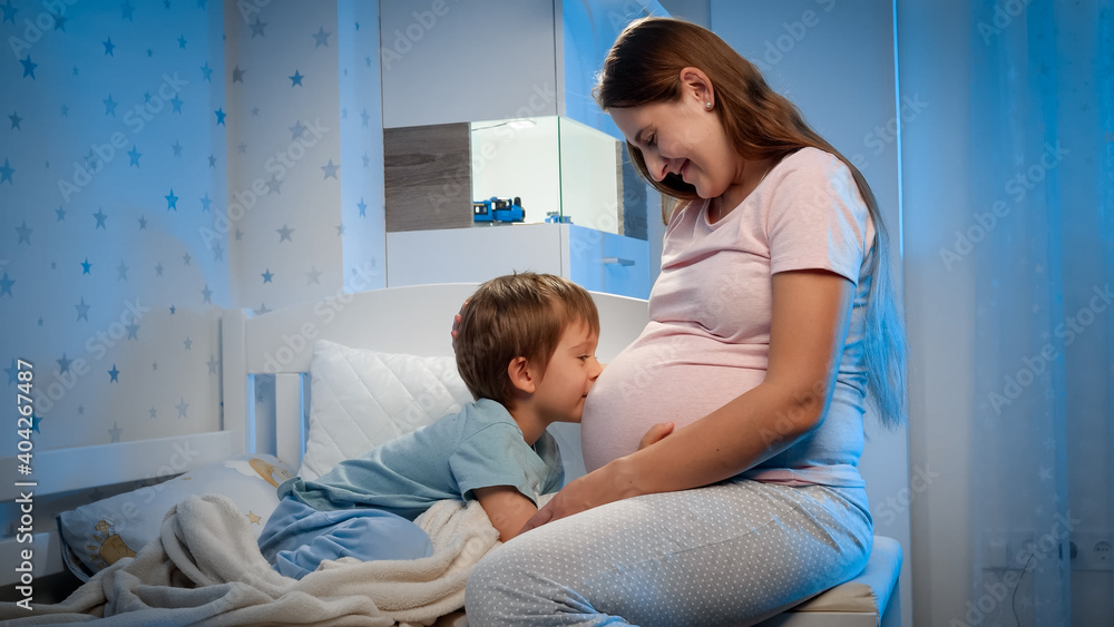 Smiling little boy hugging and talking to little unborn baby in big belly if pregnant mother sitting next to his bed at night Smiling little boy hugging and talking to little unborn baby in big belly if pregnant mother sitting next to his bed at night