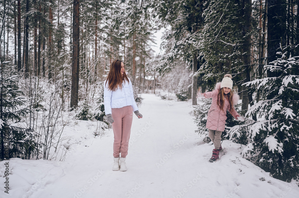 Happy mother and little cute girl in pink warm outwear walking playing snowball fight having fun in snowy white winter coniferous forest with spruce woods outdoors. Family sport vacation activities.