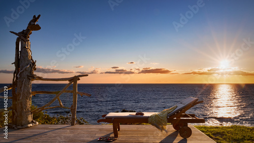 Deckchair with book, sandals and scarf on a wooden terrace overlooking the sea at sunset or sunrise