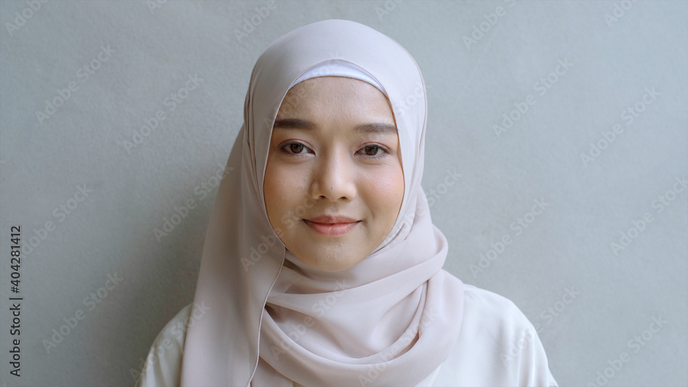 Close Up Portrait of a Young Asian Muslim Woman dressing in the traditional Hijab looking confidently and smiling at the camera with the white wall as the background Close Up Portrait of a Young Asian Muslim Woman dressing in the traditional Hijab looking confidently and smiling at the camera with the white wall as the background