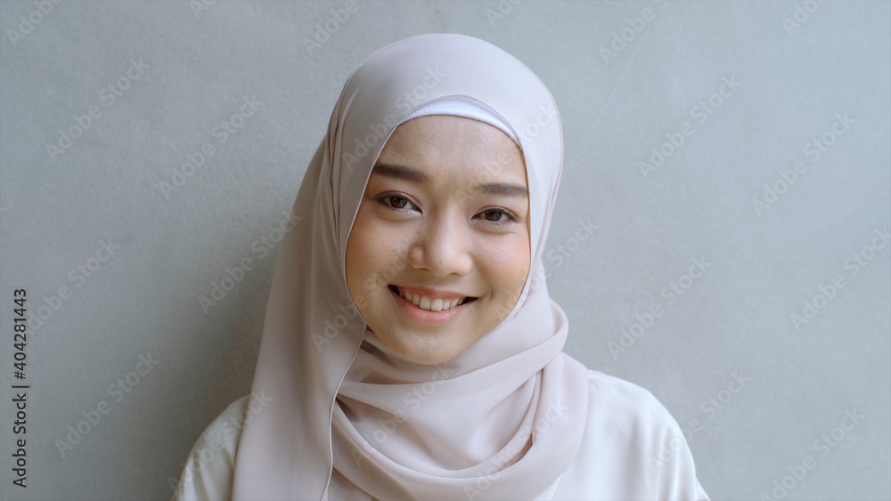 Close Up Portrait of a Young Asian Muslim Woman dressing in the traditional Hijab looking confidently and smiling at the camera with the white wall as the background Close Up Portrait of a Young Asian Muslim Woman dressing in the traditional Hijab looking confidently and smiling at the camera with the white wall as the background