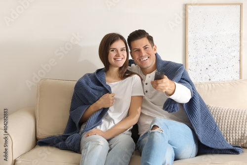 Happy couple covered with warm dark blue plaid watching television on sofa at home
