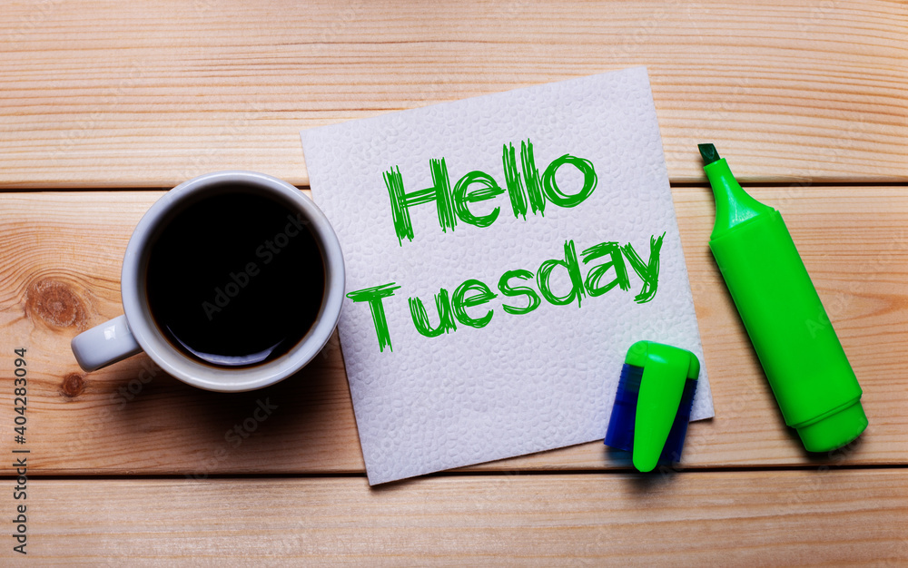 On a wooden table, a cup of coffee, a green marker and a napkin with the text HELLO TUESDAY On a wooden table, a cup of coffee, a green marker and a napkin with the text HELLO TUESDAY