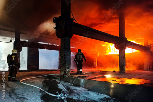 Firefighters in heat-resistant suits near an open fire. A thick layer of smoke. Behind the back of oxygen tanks On the head of protective helmets. The concept of protecting and saving the city s