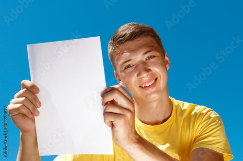 young smiling guy in a yellow t-shirt with a blank sheet of paper in his hands on a blue background
