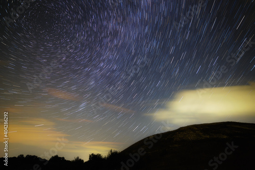 A trail of bright stars from the Milky Way in the cloudy night sky over the hills.
