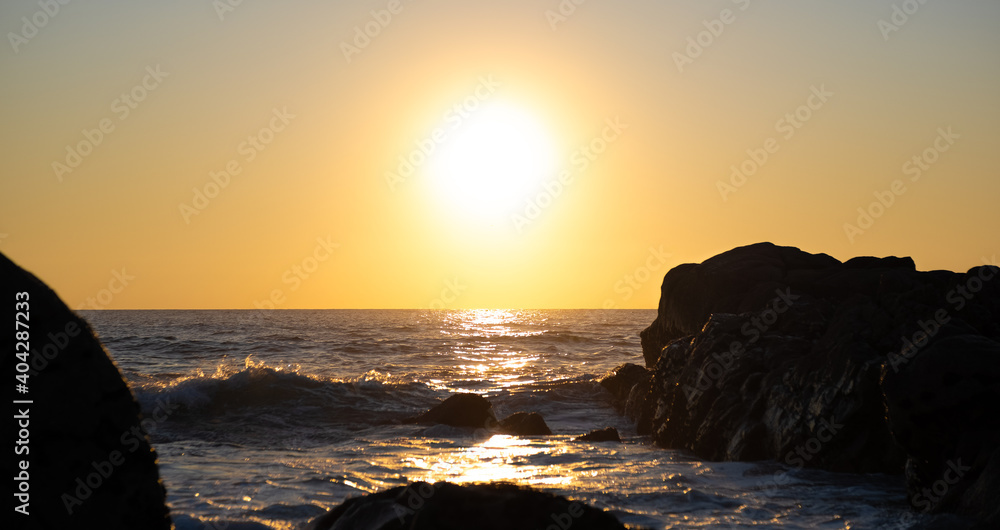 Sun setting over the ocean, with reflections on the ocean surface and dark rocks in silhouette. Shallow depth of field.