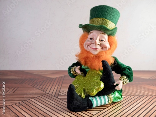 St Patrick’s Day leprechaun with a green outfit and green hat sitting on a wooden surface with a reddish orange fluffy beard, holding a glittery green four leaf clover.