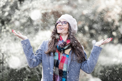 Portrait shot of attractive young woman wearing hat and winter coat while standing outside and enjoy falling snow. Happy woman standing outdoor and raising her hands while snowing heavily 