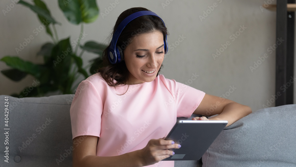 Naklejka premium Happy young indian Arabic woman sit rest on sofa at home wear headphones have video call on tablet. Smiling ethnic female in earphones use pad gadget, talk on webcam conference or watch webinar.