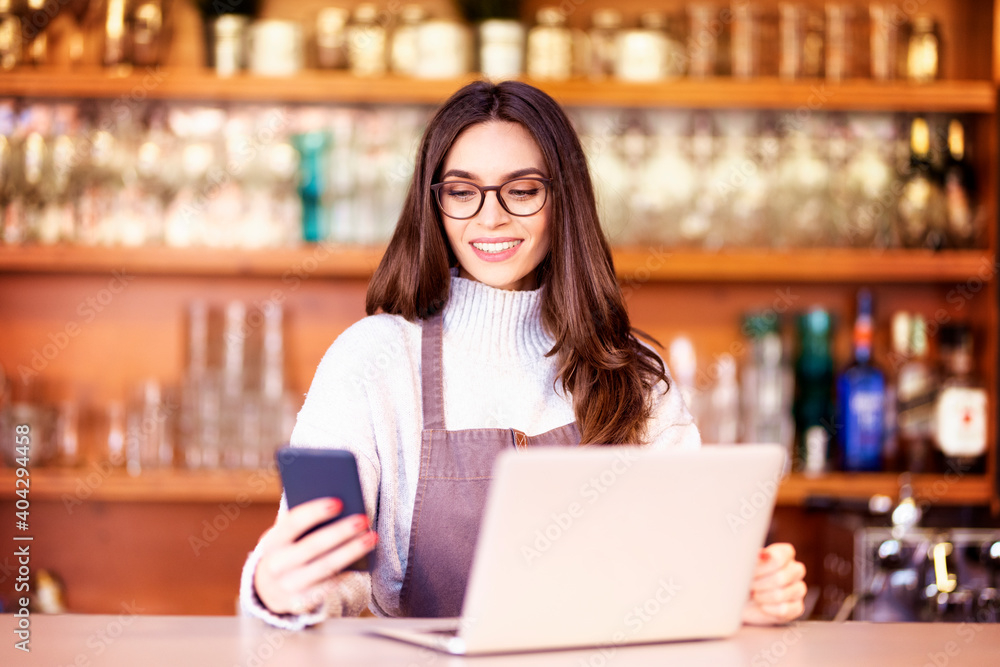 Portrait of a beautiful waitress text messaging and using laptop while standing behind the counter at a cafe. Small business. Portrait of a beautiful waitress text messaging and using laptop while standing behind the counter at a cafe. Small business.