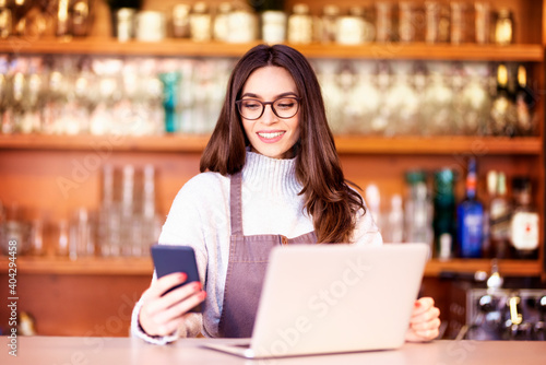Portrait of a beautiful waitress text messaging and using laptop while standing behind the counter at a cafe. Small business. 