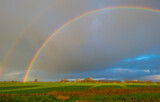 Double rainbow and yellow grey rain clouds over a windy rainy green meadow in bright sunlight in winter, Leeuwarden, Friesland, The Netherlands, January 9, 2021