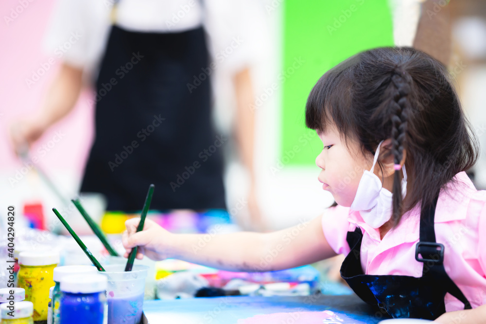 Girl is doing art in class. Child is washing the brush in a glass of water. Children take a cloth face mask on their chin. Kid is learning to paint in water. Baby 3-4 years old.