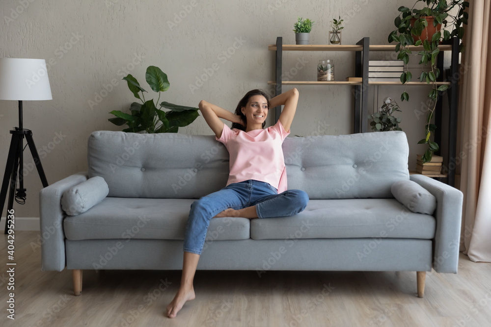 Happy millennial indian Arabic woman sit rest on comfortable couch in modern living room look in distance dreaming. Smiling young ethnic female renter or tenant relax on sofa at home, enjoy weekend.