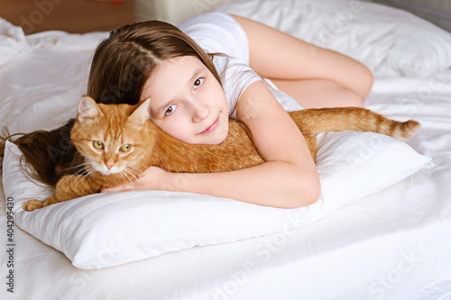A girl with a ginger cat lies on a white bed. A young girl is stroking a ginger cat while lying on a white sheet.
