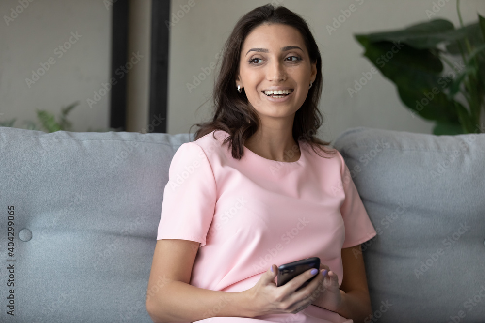 Smiling millennial indian Arabic woman relax on couch at home look in distance dreaming using smartphone. Happy young ethnic female client use browse modern cellphone think or plan, rest on sofa. Smiling millennial indian Arabic woman relax on couch at home look in distance dreaming using smartphone. Happy young ethnic female client use browse modern cellphone think or plan, rest on sofa.