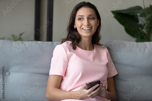 Smiling millennial indian Arabic woman relax on couch at home look in distance dreaming using smartphone. Happy young ethnic female client use browse modern cellphone think or plan, rest on sofa.