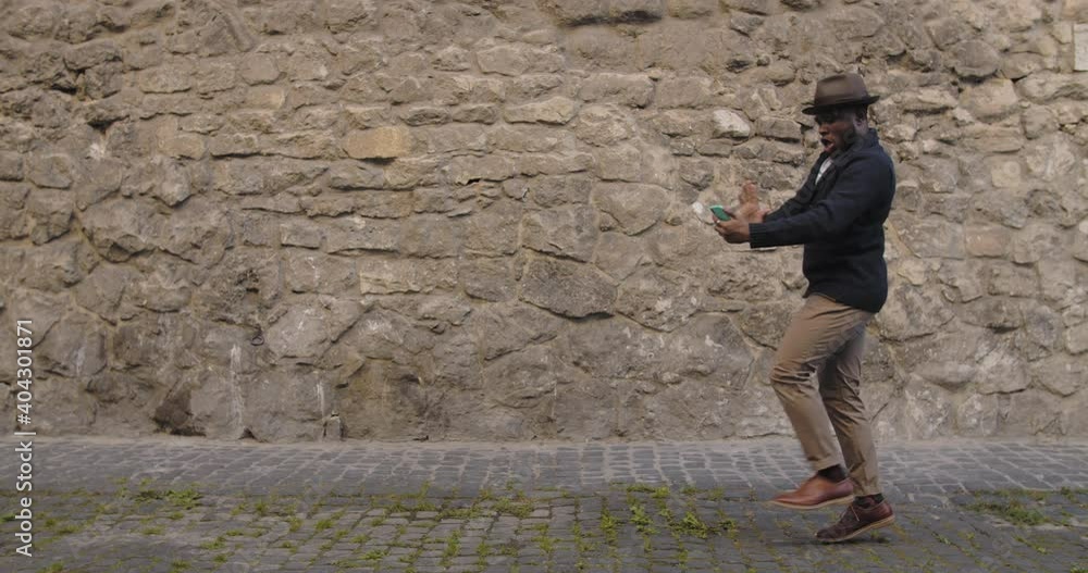 Handsome man in hat dancing and singing while looking at smartphone screen. Afro american man in trendy clothes listening to music and moving in rhytm while walking city street Handsome man in hat dancing and singing while looking at smartphone screen. Afro american man in trendy clothes listening to music and moving in rhytm while walking city street