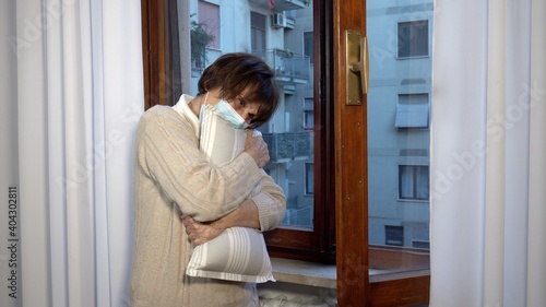 sad and lonely elderly lady woman 70 years old with mask in the apartment during Covid-19 Coronavirus lockdown quarantine home, clutching a pillow to her chest in a moment of despair