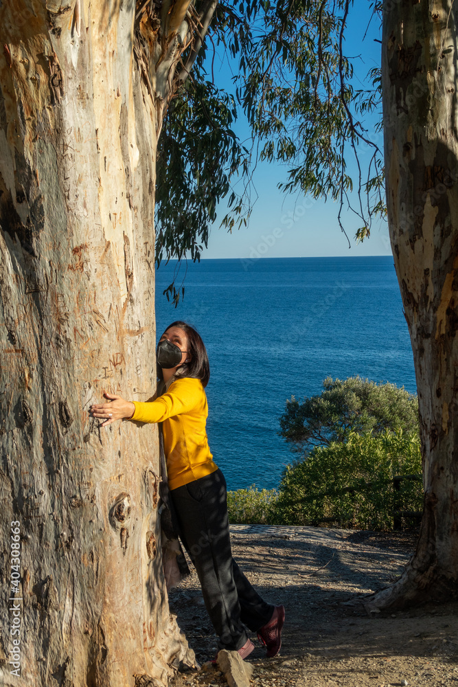 Woman with black mask hugging the trunk of a large eucalyptus with the sea in the background