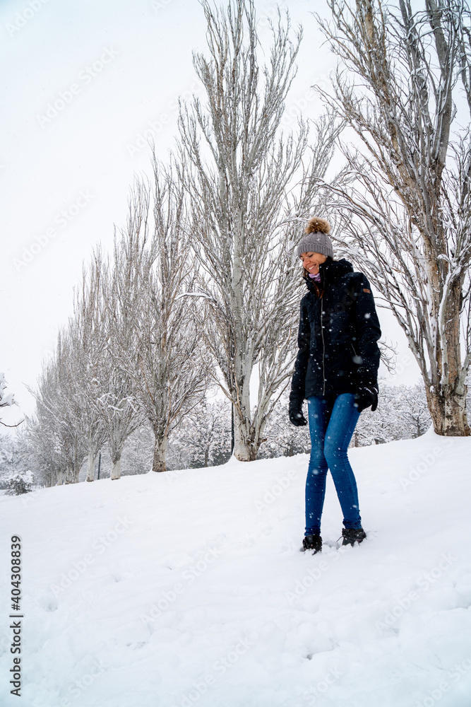 Chica joven en un parque nevado en un día de invierno con gorro de lana gris, jeans y cazadora negra