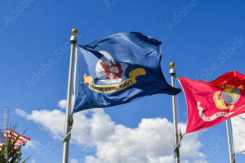 A flag of the United States Navy flies alongside and American flag and a U.S. Marine flag for the armed services in front of a blue sky.