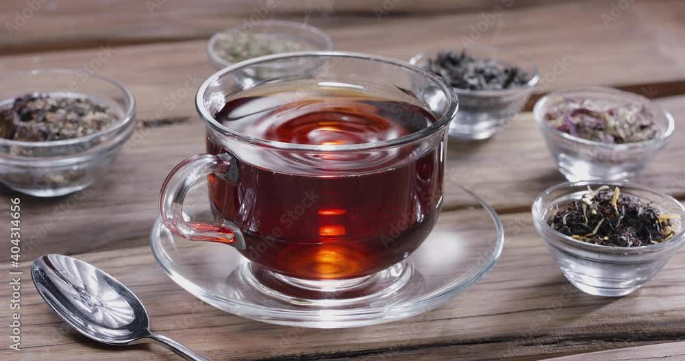 Drop slowly falls into a glass cup of tea. Around there are small bowls with different types of leaf tea. Vintage wooden table. Drop slowly falls into a glass cup of tea. Around there are small bowls with different types of leaf tea. Vintage wooden table.