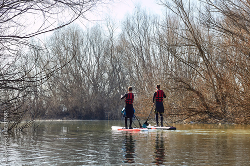 Two boys rowing on SUP (stand up paddle board) in autumn Danube river against the trees on the shore of the river