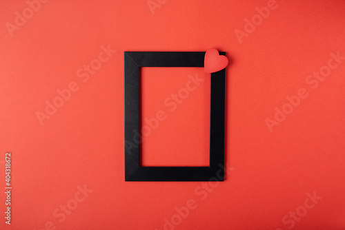 Black photo frame with red wooden heart on the red background. Valentine s Day concept. Flat lay  top view  space for text.