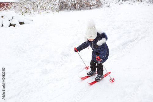 Children's feet in red plastic skis with sticks go through the snow from a slide-a winter sport, family entertainment in the open air. A little girl glides down the slope from an early age. Copy space © Ольга Симонова