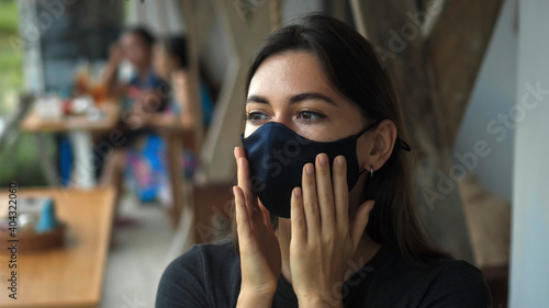 European brunette girl sits at a table in a public place and touches a soft black protective mask with her hands on her face