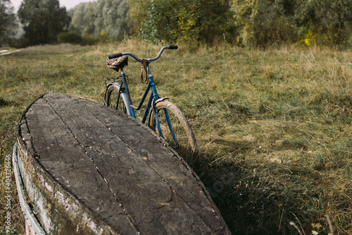 Fotografie Old rusty worn bicycle standing near weathered fishing wooden boat laying on ground with its bottom up
