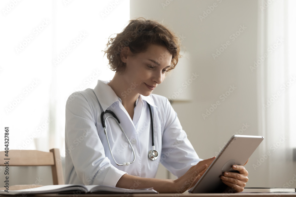 Pleasant happy female doctor general practitioner using digital computer tablet, analyzing patient's medical history or anamnesis online, giving consultation or browsing information in internet. - obrazy, fototapety, plakaty 