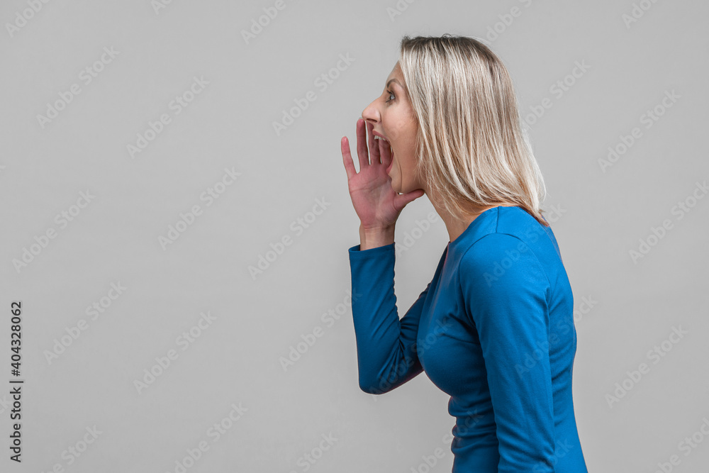 Attention! side view of aggressive woman in elegant blue dress holding her hands near wide open mouth and shouting announcement, crazy furious face. indoor studio shot isolated on gray background Attention! side view of aggressive woman in elegant blue dress holding her hands near wide open mouth and shouting announcement, crazy furious face. indoor studio shot isolated on gray background
