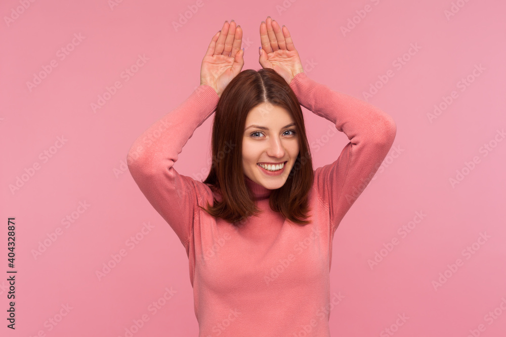 Happy playful woman with brown hair in pink sweater holding hands above head showing bunny ears, flirting, easter symbol, masquerade. Indoor studio shot isolated on pink background Happy playful woman with brown hair in pink sweater holding hands above head showing bunny ears, flirting, easter symbol, masquerade. Indoor studio shot isolated on pink background