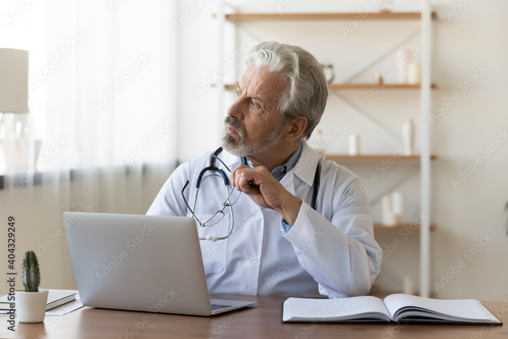 Thoughtful senior older male general practitioner looking in distance, distracted from work in hospital. Pensive middle aged doctor thinking of medical problem solution or disease treatment indoors. Thoughtful senior older male general practitioner looking in distance, distracted from work in hospital. Pensive middle aged doctor thinking of medical problem solution or disease treatment indoors.