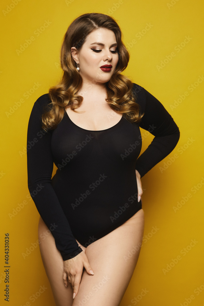 Fototapeta premium Sexy plump woman in black bodysuit posing over yellow background. Plus size model girl with bright makeup and stylish hairstyle on the yellow background, isolated