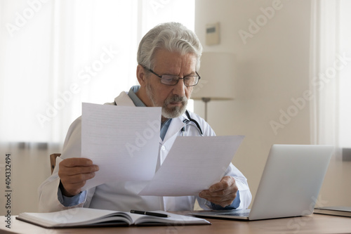 Focused middle aged senior male general practitioner in eyewear looking through paper documents, reading medical insurance contracts, reviewing reports or illness treatment history at clinic. photo