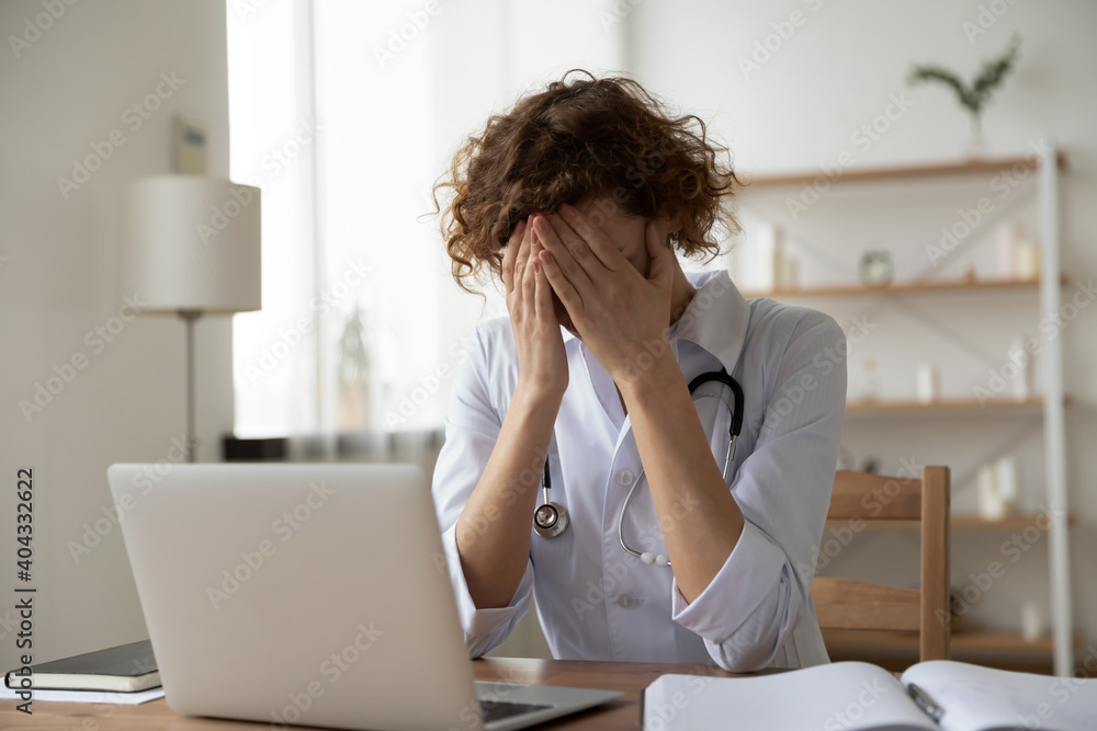 Anxious young female physician doctor covering face with hands, feeling nervous or exhausted at clinic office. Unhappy 35s general practitioner suffering from severe headache at hospital workplace. Anxious young female physician doctor covering face with hands, feeling nervous or exhausted at clinic office. Unhappy 35s general practitioner suffering from severe headache at hospital workplace.