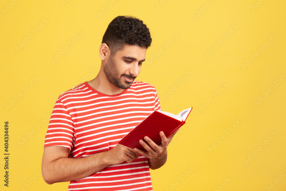 Concentrated bearded man holding book and smiling, enjoying reading, standing absorbed with exciting plot, checking schedule in notepad. Indoor studio shot isolated on yellow background Concentrated bearded man holding book and smiling, enjoying reading, standing absorbed with exciting plot, checking schedule in notepad. Indoor studio shot isolated on yellow background