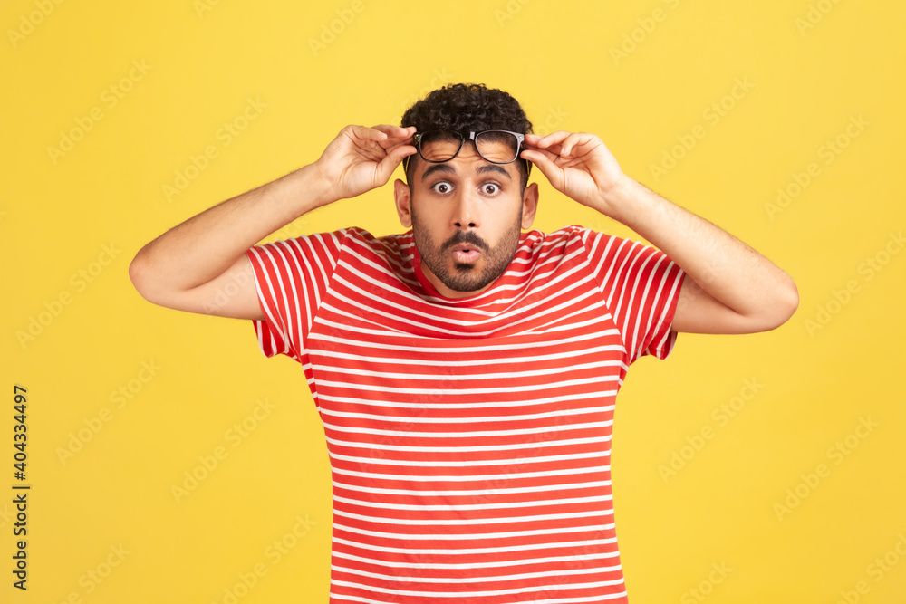 Overwhelmed bemused man with beard in striped t-shirt raising eyeglasses and looking at camera with big eyes and shocked surprised expression. Indoor studio shot isolated on yellow background Overwhelmed bemused man with beard in striped t-shirt raising eyeglasses and looking at camera with big eyes and shocked surprised expression. Indoor studio shot isolated on yellow background