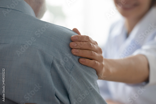 Close up young kind happy female doctor putting hand on old pensioner's shoulder, giving psychological support to elderly man at checkup meeting in hospital, showing care, medical service concept.