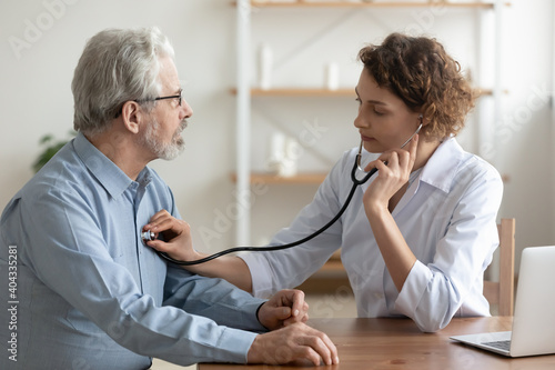 Focused young female cardiologist or general practitioner in white coat listening heartbeat or elderly senior male patient at checkup meeting, preventing cardiovascular disease, healthcare concept. photo