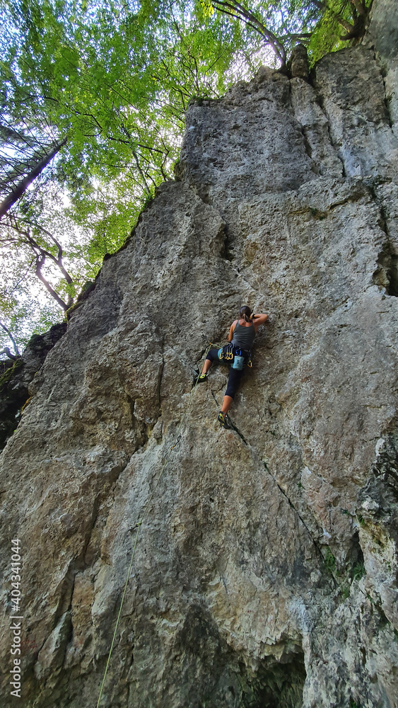VERTICAL: Young athletic woman top rope climbs up challenging cliff in Slovenia.