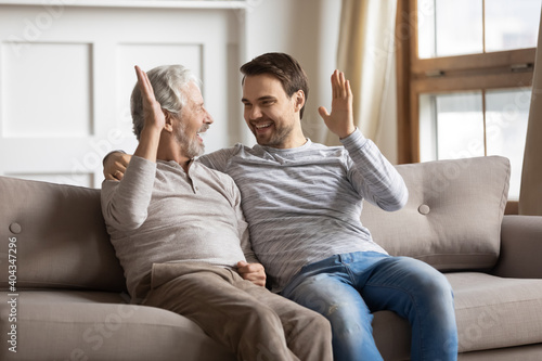 Excited millennial Caucasian man and senior father relax at home give high five make deal. Smiling elderly dad and adult grownup son sit on sofa celebrate win or victory. Family bonding concept.