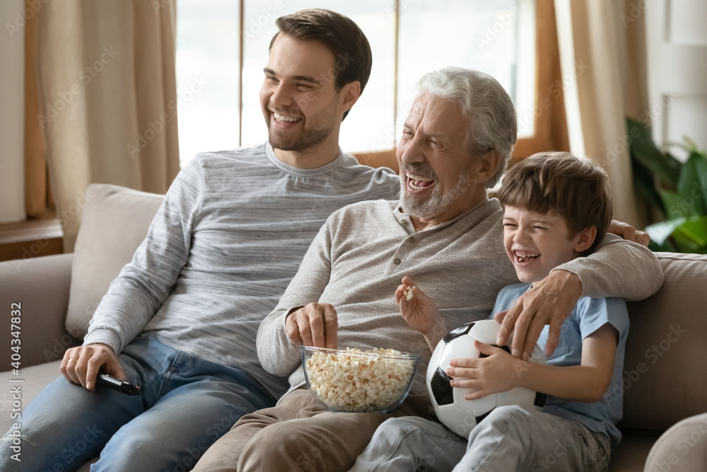 Overjoyed three generations of men relax on sofa in living room watch football match eat popcorn. Happy boy with young father and elderly grandfather have fun rest at home on family weekend together. Overjoyed three generations of men relax on sofa in living room watch football match eat popcorn. Happy boy with young father and elderly grandfather have fun rest at home on family weekend together.