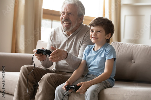 Obraz na plátně Overjoyed elderly Caucasian grandfather have fun play video computer games with small grandson at home