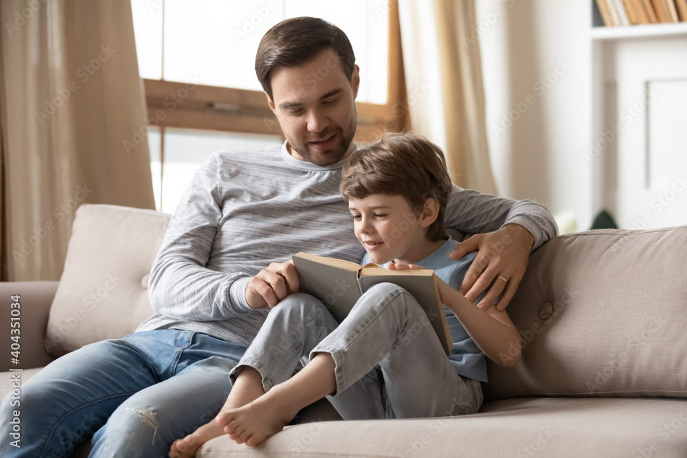 Happy young Caucasian dad and little son sit rest on sofa in living room read book at home together. Loving caring father teach small boy child learn prepare for school. Children education concept.